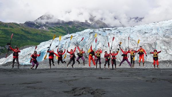 14 NOLS students holding kayak paddles jump for joy in front of an Alaskan glacier