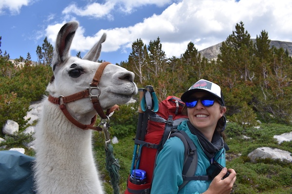 Smiling NOLS student backpacks beside a pack llama in the Wind River Range