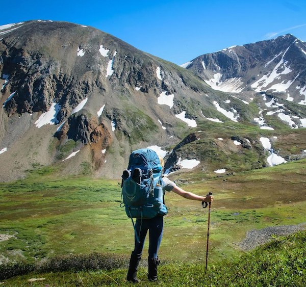 NOLS student with backpack and trekking pole looks across a green valley toward snow-dotted mountains