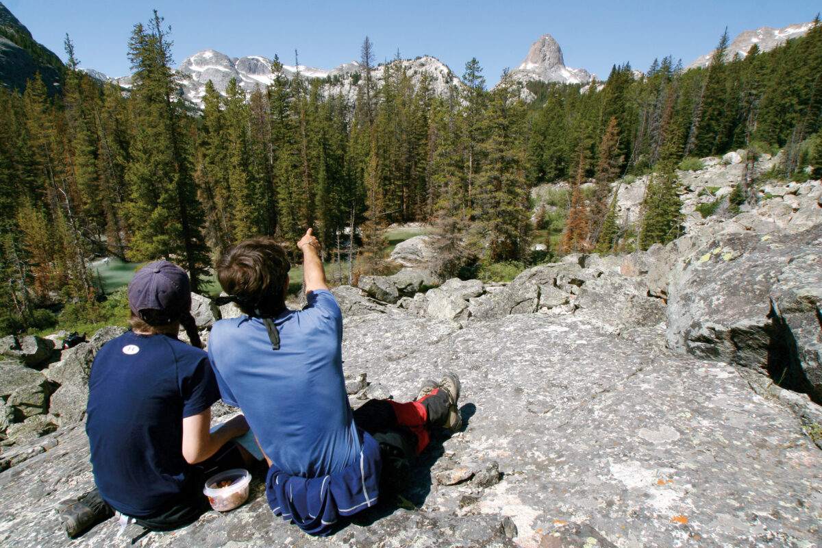 two teens facing away sitting on a rock, the one on the right pointing towards something far off into the trees in front of them