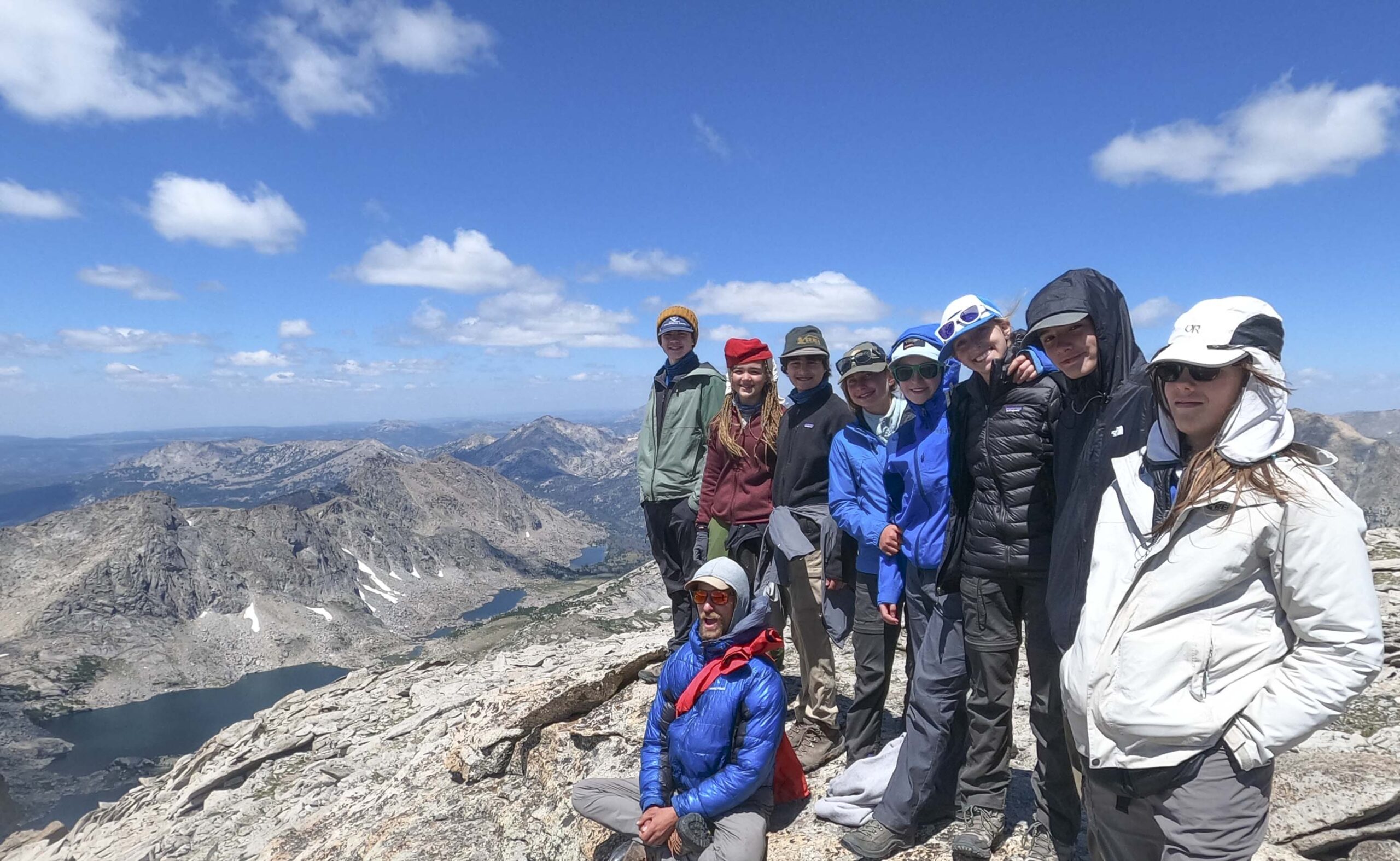 Group of teens standing on top of a large rock at elevation with mountains off in the background