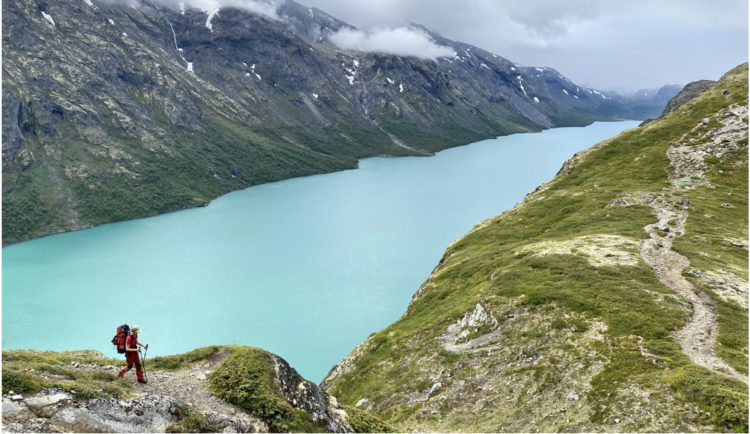 A person hikes in the foreground. Behind them is a turquoise lake nested between two steep rocky mountains.