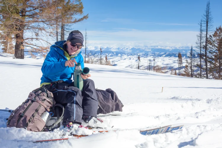 a woman sits atop a backpack on the snow. She's laughing and pouring something from a Thermos.