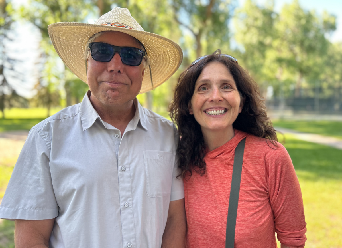 Andy Blair, wearing a sunhat, and Jacki Klancher stand close together with big smiles on their faces. Green grass and green deciduous trees in the background.