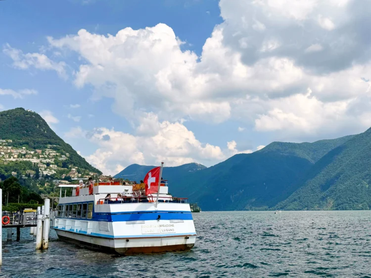 A blue and while boat with a Swiss flag arrives to the dock on the lake. Mountains and buildings rise up in the distance.