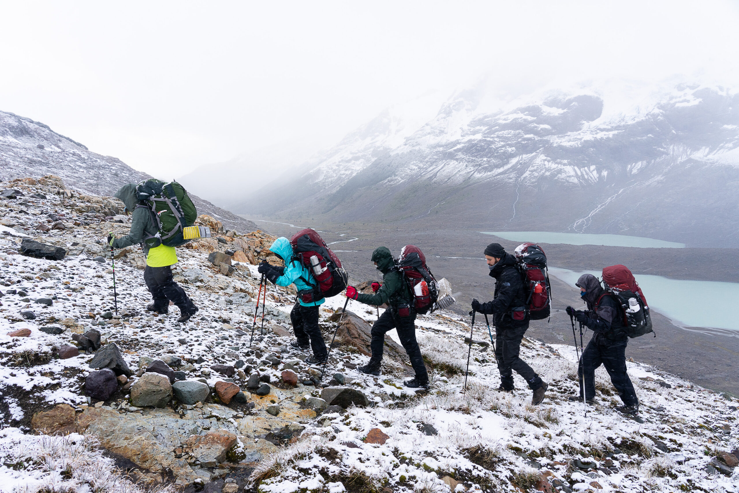 a group of people wearing backpacks hike up a hillside during a snowstorm