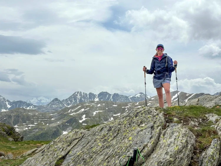 Hikers smiles and hold trekking poles with mountains behind her.