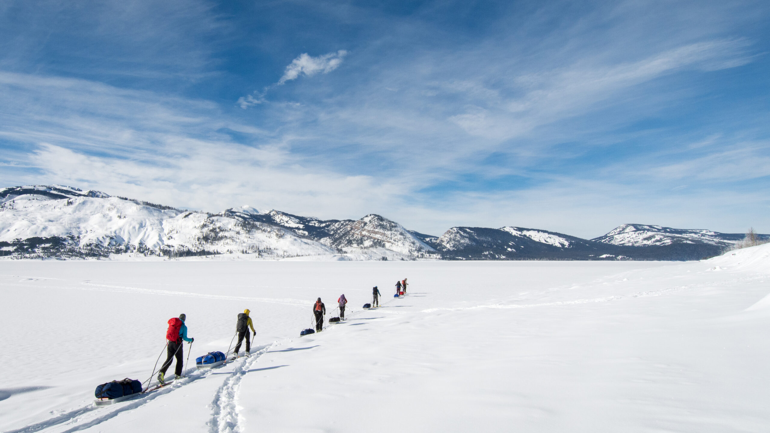 A line of people ski across an open expanse of snow. there are tall mountains in the background. the sky is blue.