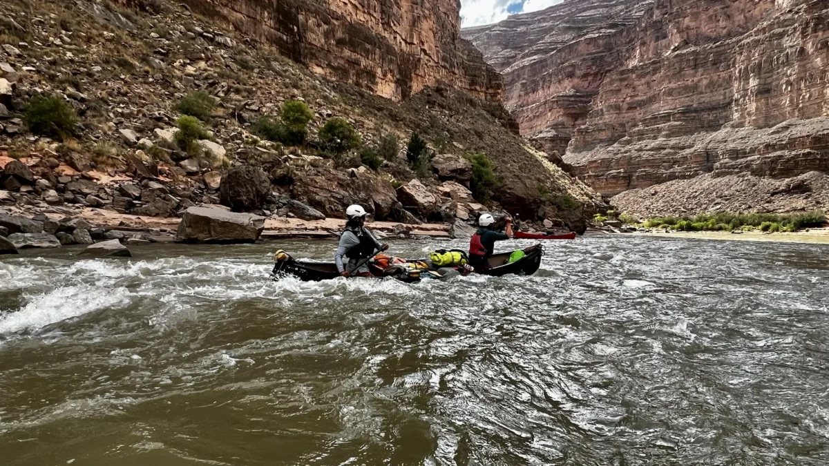 two people wearing helmet and pfds paddle a canoe along a canyon wall