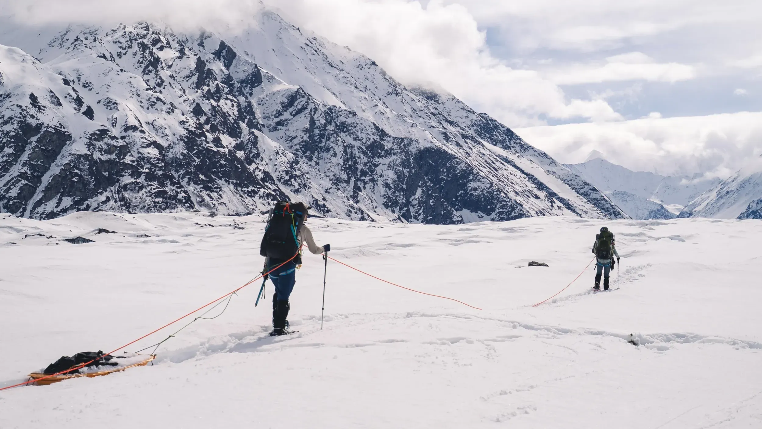 two people on a rope team haul sleds across a snowy expanse. there are mountains in the background