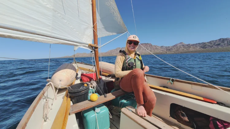 a woman in a baseball hat and sunglasses sits in a small sailboat, smiling at the camera